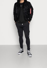 Giacca bomber nera con zipper, t-shirt nera e pantaloni cargo neri. Abbinata a sneakers bianche e nere. Tessuto liscio, tasche funzionali.