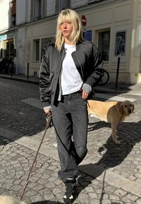 Chaqueta bomber negra, camiseta blanca, vaqueros negros y zapatillas negras. Pelo rubio corto, sosteniendo la correa de un perro en una calle empedrada.