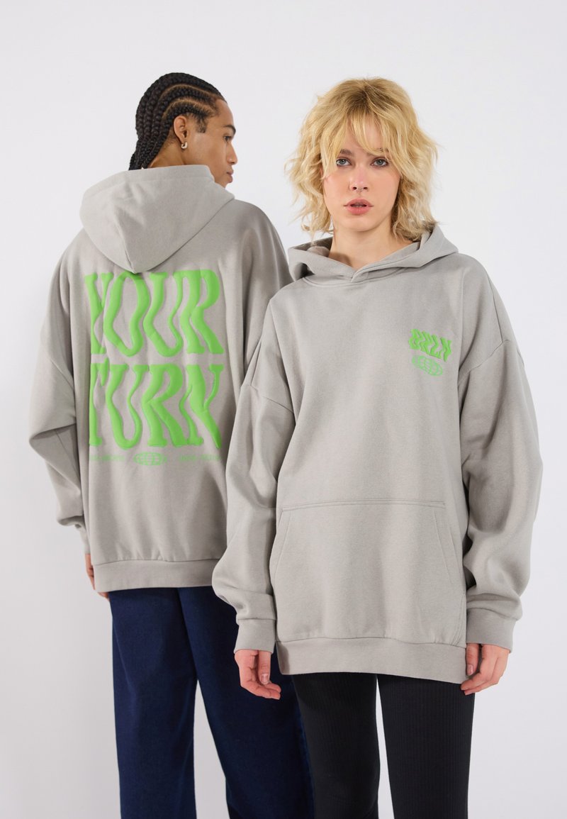 Sweatshirts gris surdimensionnés avec une poche avant, présentant un grand texte vert "À VOTRE TOUR" au dos et un petit logo en bas à gauche.