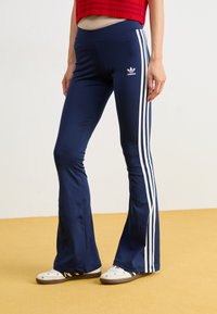 Sötétkék, bővülő sportnadrág fehér háromcsíkos díszítéssel az oldalán; sima, rugalmas anyagból készült, Adidas logóval.