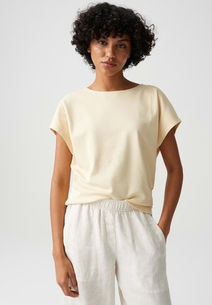Femme aux cheveux bouclés portant un top beige à manches courtes et un pantalon clair à taille élastique, debout devant un fond uni.