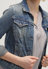 Denimjakke i en gradient blå vask, med to brystlommer, sølvfarvede knapper og opkrøllede ærmer over en lyse mønstret top.