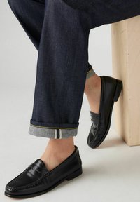 Czarne skórzane mokasyny z haftowanym wzorem i niskim obcasem, noszone z ciemnymi dżinsami z podwiniętymi mankietami i kontrastującym przeszyciem.