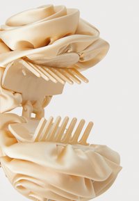 Clips de cabelo em bege claro com um design em tecido acetinado e detalhes plissados, apresentando dentes semelhantes a um pente para uma fixação segura.