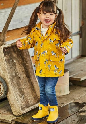Impermeable amarillo con patrón de cebra y bolsillos frontales, combinado con botas de lluvia amarillas. Jeans azules. Niño sonriendo, de pie sobre un suelo de madera.