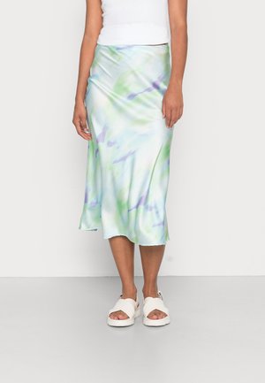 Femme portant une jupe midi tie-dye pastel avec des nuances de vert, bleu et violet, associée à des sandales blanches et un top blanc sans manches.