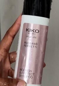 Schampoflaska med en blank svart kork och en vit botten, med en ljusrosa etikett med svart text. Texturen ser våt ut med vattendroppar.