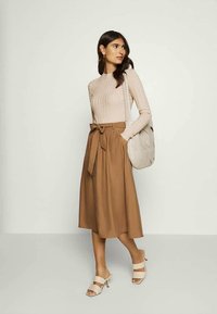 Beige getextureerde langwerpige top gecombineerd met een bruine midi-rok met een strikband aan de voorkant. Gecombineerd met een lichtgrijze tas en lichte hakken.
