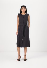 Vestido cinza escuro sem mangas com fecho de botão à frente, faixa na cintura e ombros com aba. Combinado com sandálias de salto alto pretas com padrões de bolinhas.