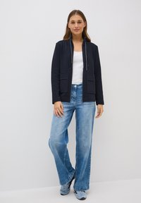 Dunkelblaue Zip-Jacke mit hohem Kragen, zwei Fronttaschen; kombiniert mit hellblauen weit geschnittenen Jeans und grauen Sneakers. Glatte Textur.