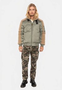 Olivengrøn pufferjakke med ærmer i beige fleece, lynlås foran og sidelommer. Kombinationen består af camouflage cargobukser og sorte snørestøvler.