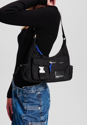 Femme portant un pull noir côtelé et un jean bleu tenant un sac à bandoulière noir Karl Lagerfeld avec des tirettes de fermeture éclair bleues et une poche avant à boucle.