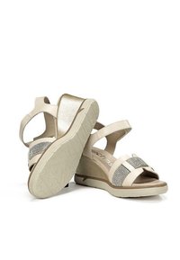 Sandalias de cuña beige con una correa en el tobillo, acentos metálicos y una suela texturizada. Cuenta con detalles de strass en la correa delantera.