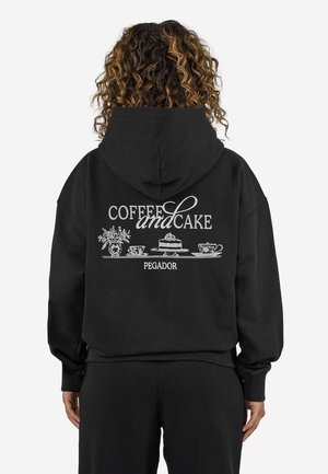 Person mit lockigem Haar, die einen schwarzen Hoodie mit dem Text "Coffee and Cake" und Illustrationen von Kuchen, Tassen und Blumen auf dem Rücken trägt.