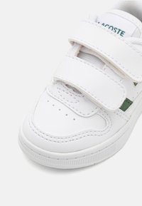 Scarpa bianca con fascia in Velcro, tomaia in pelle texture, area della punta perforata e dettaglio verde vicino al tallone. Punta arrotondata e suola in gomma.