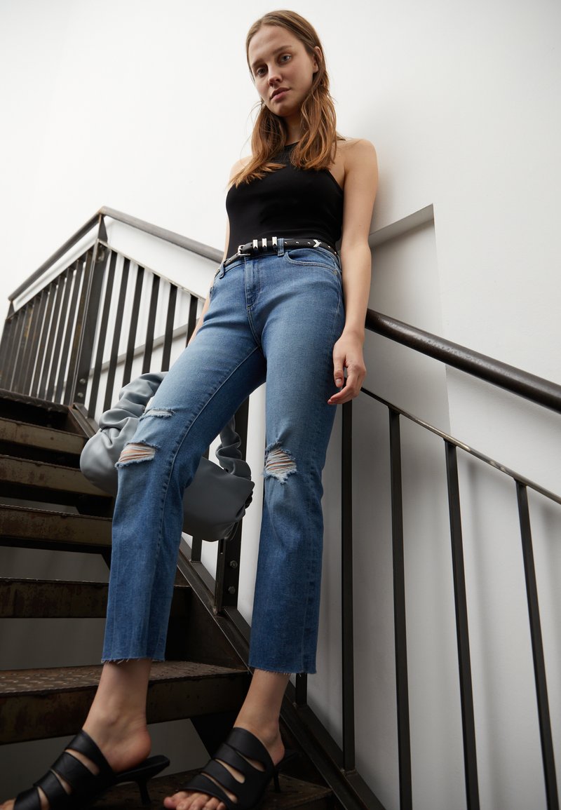 Jeans rasgados azul claro com corte reto, cinto preto e sandálias pretas com tiras. A modelo segura uma bolsa cinza descontraída enquanto está numa escada.
