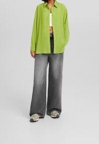 Camicia verde a maniche lunghe con texture sopra una maglietta bianca corta, abbinata a pantaloni in denim grigi larghi e sneakers grigie, creando un look a strati.