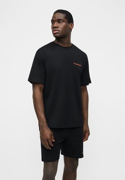 Άνδρας που φοράει μαύρο κοντομάνικο μπλουζάκι με μικρό πορτοκαλί κείμενο και μαύρο σορτς, στέκεται μπροστά σε απλό λευκό φόντο, κοιτάζοντας προς τα αριστερά.