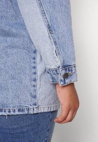 Dłoń osoby oparta obok rękawa jasnoniebieskiej kurtki dżinsowej z zapięciem na guzik, w połączeniu z ciemniejszymi niebieskimi spodniami dżinsowymi.