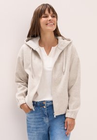 Haut de sweat à capuche zippé gris clair avec une texture douce, présentant une doublure intérieure blanche et des cordons de serrage, associé à un haut blanc et un jean bleu.