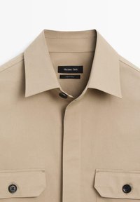 Camicia beige con colletto a punta, due tasche sul petto, bottoni neri e tessuto liscio. Etichetta con la scritta "Massimo Dutti, made in Turkey."