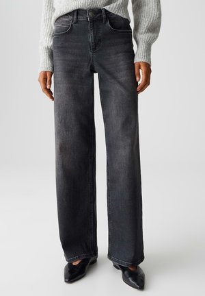 Persoon die een zwarte wijde broek met hoge taille draagt, een grijze gebreide trui en zwarte puntige schoenen, staand tegen een effen achtergrond.