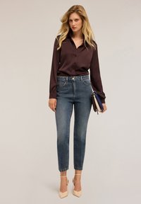 Blonde vrouw in een donk paars knoopblouse, blauwe hoge taille jeans, beige hakken, met drie kleine tasje in haar handen.