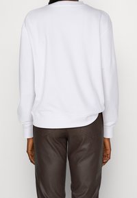 Sudadera blanca con cuello redondo, hombros caídos y dobladillo curvo, combinada con pantalones marrones texturizados. Diseño simple, tela suave.