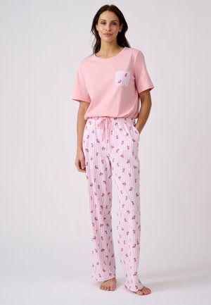 Vrouw staat op blote voeten, draagt een roze korte mouwen top met gestreepte bloemenzak en bijpassende bloemenpyjamabroek met trekkoord.