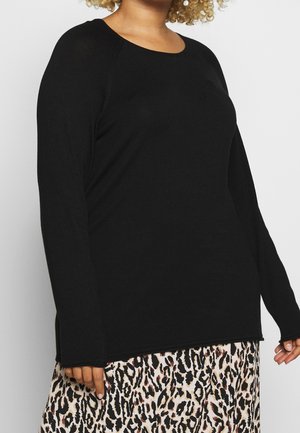 Personne portant un haut noir à manches longues associé à une jupe beige et noire à imprimé léopard sur un fond clair uni.