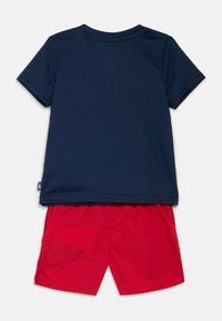 Granatowy T-shirt z krótkim rękawem i okrągłym dekoltem, zestawiony z czerwonymi szortami. Materiał T-shirtu jest gładki; szorty są lekkie i lekko teksturowane.