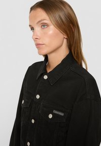 Melns denima jaku ar sudraba pogām, plakanu apkakli un divām krūšu kabatām. Uz vienas kabatas ir neliels Calvin Klein logo.