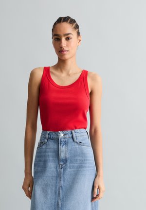 Femme aux cheveux tressés portant un haut rouge sans manches et une jupe en denim bleu clair taille haute, debout devant un fond gris clair uni.