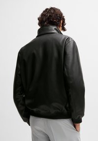 Svart kunstlærjakke med høy krage, rynket kant og mansjetter; har en glatt tekstur og løs passform.