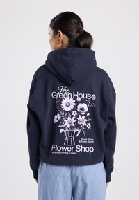 Sweat à capuche bleu marine avec un grand motif floral rose au dos, représentant une cafetière et le texte "Green House Flower Shop".