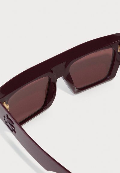 Lunettes de soleil bordeaux avec des montures géométriques, verres teintés foncés et accents dorés sur les branches. Matériau en plastique lisse et brillant.