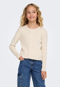 Krämfärgad ribbad kofta med knappstängning, långa ärmar och kort design, kombinerad med blå cargo-jeans. Mjuk textur och klassisk stil.