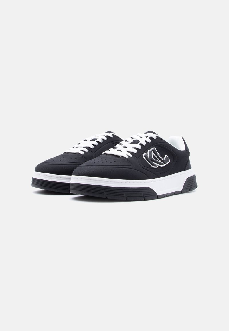 Zapatillas negras de perfil bajo con cordones blancos, suelas blancas gruesas y un logotipo estilizado en forma de "K" en el lateral, sobre un fondo blanco liso.