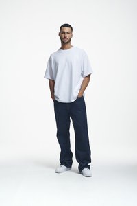 Weißes, übergroßes T-Shirt aus Baumwolle, kombiniert mit dunkelblauen, lockeren Jeans und weißen Sneakern, vor einem schlichten Hintergrund stehend.