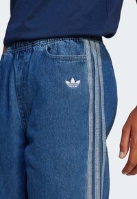 Τζόγκες από τζην σε μεσαίο μπλε χρώμα με ελαστική μέση, διαθέτοντας λευκό λογότυπο Adidas και γκρι τρεις λωρίδες κατά μήκος των πλευρών.