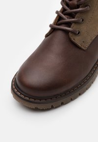Botte en cuir marron avec des accents en daim beige, bout rond et semelle épaisse en caoutchouc. Présente des lacets ronds et des coutures contrastées.