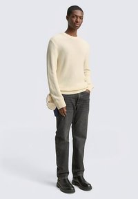 Krēmkrāsas ribotais džemperis ar garām piedurknēm, kombinēts ar tumšām džinsa biksēm un melniem potīšu zābakiem; rokās tur mazu bēša krāsas somiņu.