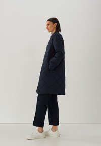 Marineblauwe gewatteerde lange jas met een zak, gekenmerkt door een rechte snit en een vlakke textuur, gecombineerd met donkere broeken en witte sneakers.