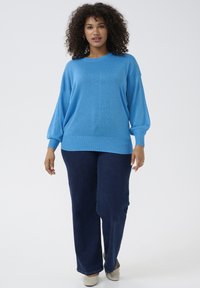 Lichtblauwe, geweven oversized trui met lange, opgeblazen mouwen. Gepaard met donkerblauwe, rechte jeans en beige schoenen.