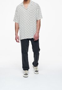 Camicia bianca a maniche corte con colletto e motivo geometrico nero, abbinata a jeans grigio scuro e sneakers grigie chiare.