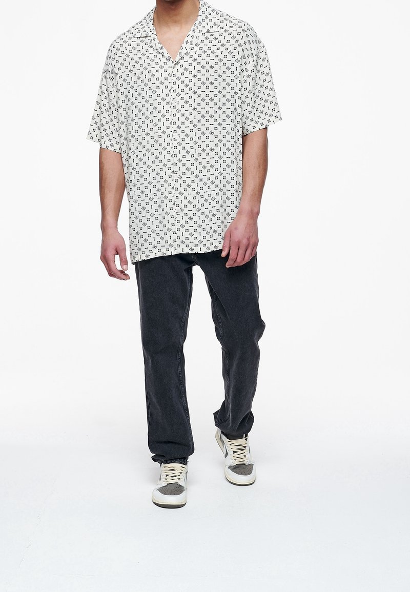 Camicia bianca a maniche corte con colletto e motivo geometrico nero, abbinata a jeans grigio scuro e sneakers grigie chiare.