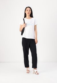 White cotton t-shirt with blue text, black drawstring pants, and white block-heeled sandals. Model holding a black bag.