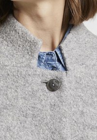 Grå texturerad kappa med hög krage, med en rund, mörk knapp och en denimskjorta under. Tyget verkar mjukt och tjockt.