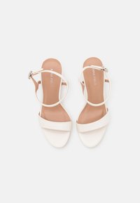 Sandales à talons hauts blanches avec une texture lisse, une bande de cheville ajustable et une large bande à l'avant. Design minimaliste avec un détail matériel subtil.