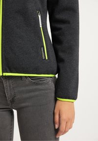 Mörkgrå zip-up jacka med neongröna detaljer, tillverkad av mjukt tyg. Har en sidoficka och en figursydd design vid midjan.
